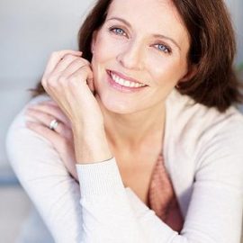 A smiling woman with brown hair, resting her chin on her hand, wearing a white sweater and a pink top.