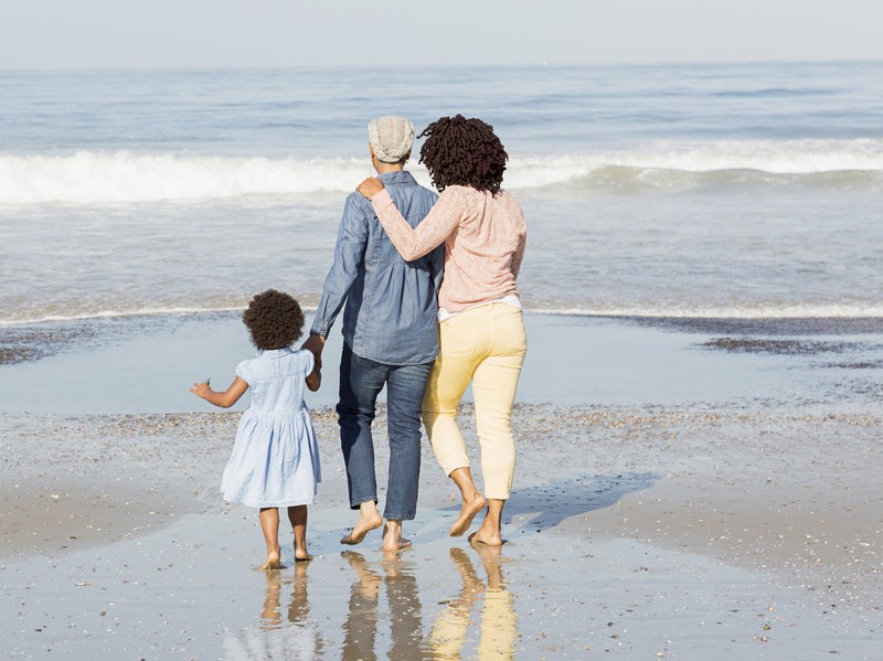 A family of three is walking hand-in-hand along the shore, with their backs to the camera, facing the sea.