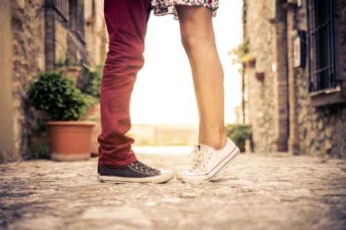 Two people standing on a cobblestone street touching toes with a blurred old-town backdrop.