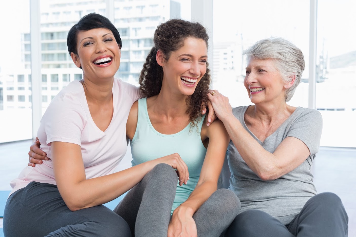Three women of varying ages sitting together, smiling and embracing in a bright room.