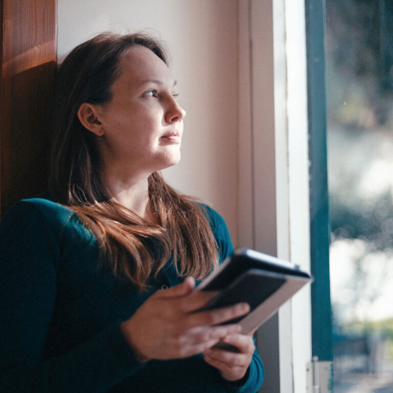 A woman holding a book gazes pensively out a window with soft light on her face.